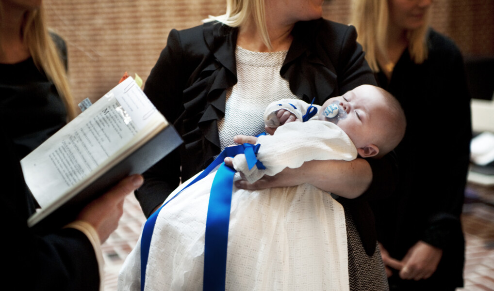 Mor holder dåbsbarn i hvid dåbskjole, mens præst læser fra dåbsritualet ved barnedåb i moderne kirke